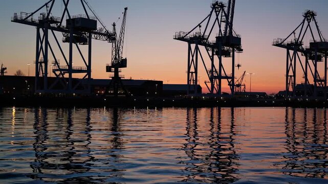 Cranes stand tall at the port as the sun sets in the background. The sky changes color and reflections shimmer on the water surface near the docks in the evening.