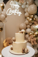 A festive two-layer cake for a holiday with a candle on top stands on a table against the background of a screen with inflated balloons and a neon inscription
