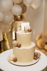 A festive two-layer cake for a holiday with a candle on top stands on a table against the background of a screen with inflated balloons