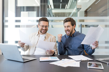 Two happy male business partners are celebrating achievement and financial success together in the modern office, looking at documents with excitement and triumph