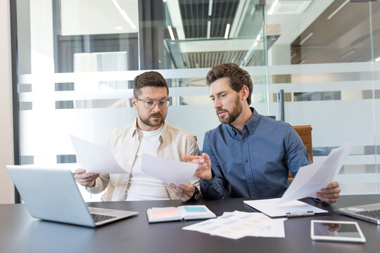Two male business professionals are reviewing documents and discussing a project, collaborating effectively at a conference table with laptops and papers in a contemporary corporate office setting