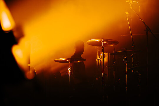 Silhouette of a drummer standing with his back to the camera in front of a drum set, illuminated by bright yellow stage lights. 