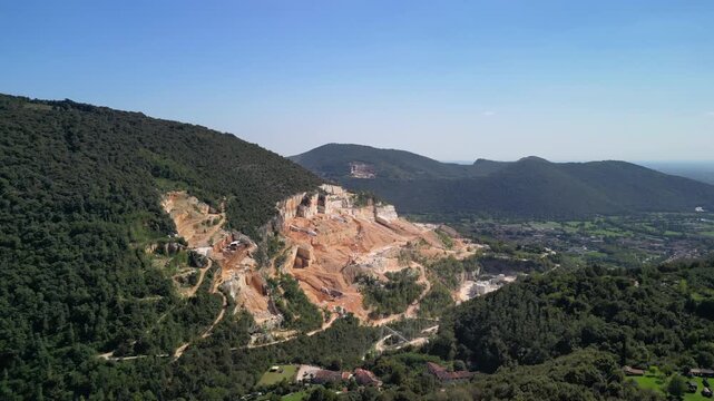 Aerial drone glide toward Botticino limestone quarry near Brescia Italy, stepped open pit walls and dusty benches contrast with forest ridges and a broad valley