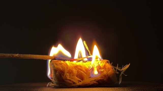 A thin wooden matchstick is held against a compact roll of shaved wood kindling. As the flame catches the dry fibers, it produces a steady, soft hissing and crackling sound.