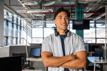 Fototapeta premium Asian man in his 20s standing with arms crossed, wearing striped shirt and headphones in office