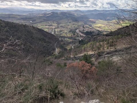 Panorama montano con una valle sinuosa che si perde tra colline e rilievi distanti. In primo piano si vedono alberi spogli e arbusti tipici della stagione invernale