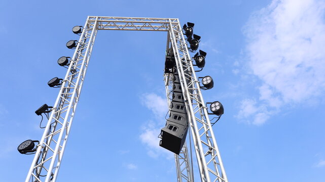 Speaker and lighting system on a metal frame. Line array speakers hanging from a pole beside an outdoor concert stage on a blue sky background with text copy space with selective focus.