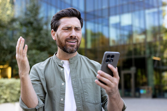 Man experiencing frustration and dissatisfaction while using his smartphone, receiving bad news, looking upset with his hand raised, standing in front of a modern office building