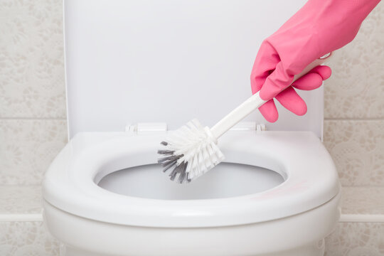 Woman hand in pink rubber protective glove holding brush and cleaning white toilet. Closeup. Front view.