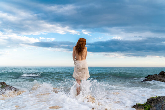 Back view of redheaded shirtless young woman standing in front of the sea