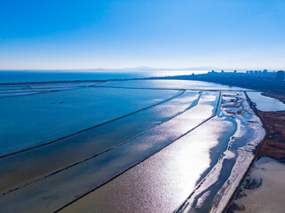 View of salt flats by the coast with water channels and city skyline in the background under clear blue sky during daytime