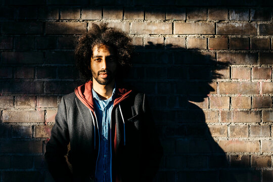 Portrait of a confident young man standing at a brick wall in shadow