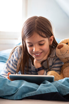 Portrait of smiling girl lying on bed with teddy bear using digital tablet