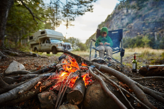 Chile, Santa Magda, Rio Maniguales, boy sitting at campfire in forest with camper van in background