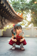 Happy little Asian girl in traditional Chinese dress holding red envelope angpao, cheerful child celebrating Lunar New Year festival at shrine, concept of fortune and luck.