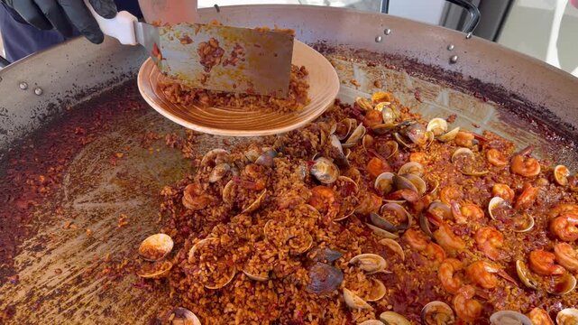 Close-up of traditional Spanish seafood paella with shrimp and clams being served from a large pan in a restaurant kitchen, showcasing Mediterranean cuisine and fresh ingredients