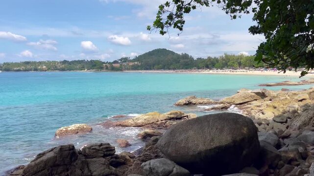 Scenic daytime view of Surin Beach in Phuket with turquoise water, rocky shoreline in the foreground, lush green hills, and a sandy tropical beach in the distance, Thailand