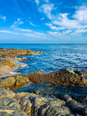 Waves crashing on rocky shore with distant boats under a blue sky on a sunny day near a coastal area