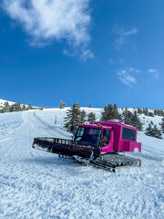 Large pink grooming machine is parked on snowy slope in a winter landscape under a bright blue sky