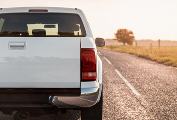 Close-up of white pickup truck rear and. Boxy 4x4 on a countryside paved road in late afternoon. Rear view of white truck against the sun, lone tree on the right. © andrbk
