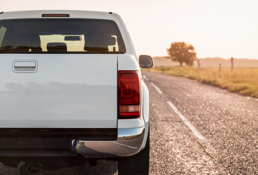 Close-up of white pickup truck rear and. Boxy 4x4 on a countryside paved road in late afternoon. Rear view of white truck against the sun, lone tree on the right.
