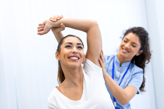 A smiling patient performs shoulder stretching exercises assisted by a female physiotherapist. Professional rehabilitation and physical therapy for arm mobility and injury recovery in a bright office.