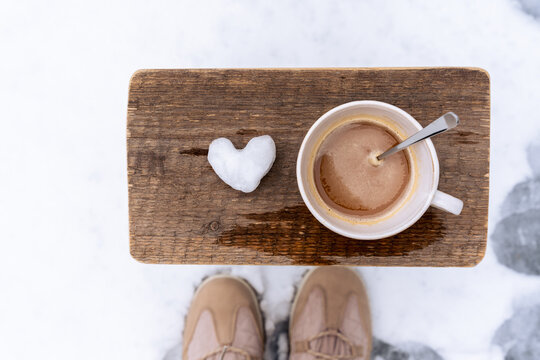 Draufsicht auf eine Tasse Kaffe und ein Herz aus Schnee auf einem Holz Hocker im Schnee. Winter, K&auml;lte.