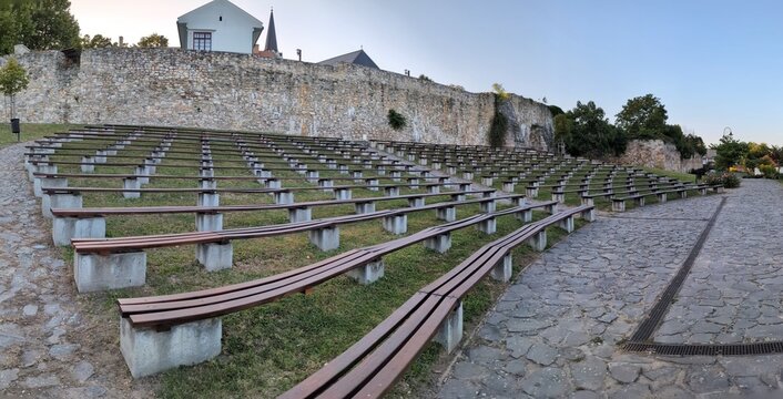 Wide-angle view of an empty amphitheater. Curved wooden benches on concrete supports, arranged on a grassy slope. Framed by historic stone walls and cobblestone paths, a church tower and greenery.