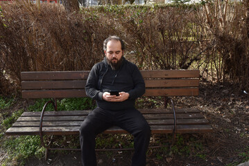 Fototapeta premium Bearded man in black casual clothing sitting on a wooden park bench while looking at his smartphone