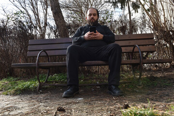 Fototapeta premium Bearded man in black casual clothing sitting on a wooden park bench while looking at his smartphone