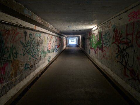 Moody, atmospheric view of a long pedestrian tunnel lined with graffiti on the walls. Perspective toward a distant exit glowing with daylight. Contrast between the dim interior and  the bright opening