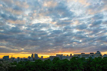 Kyiv cityscape featuring a vibrant sunrise with golden light rays peaking through a cloudy sky, illuminating the urban skyline and green trees in the foreground