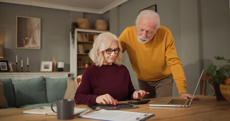 Obraz premium Elderly couple sits at a table in their living room, looking at a tablet and discussing financial planning related to life insurance. They are focused and engaged in their conversation.