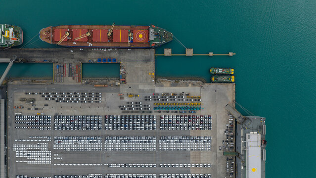 Aerial view of cargo ships docked beside rows of cars, a symphony of steel and sea against the turquoise waters, Tambon Thung Suk La, Chang Wat Chon Buri, Thailand.
