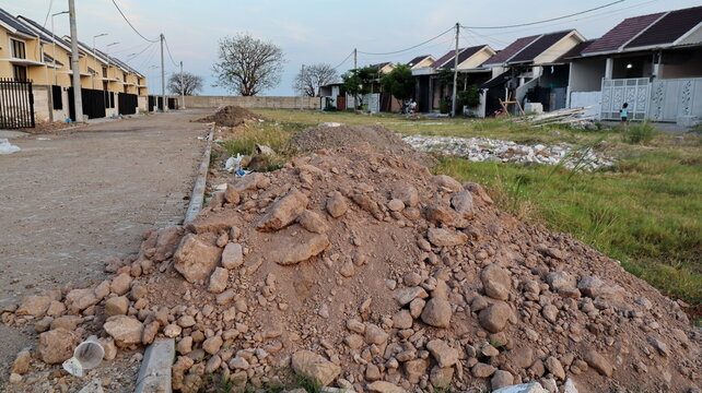 A mound of soil mixed with large rocks lies beside a paved sidewalk and grassy field, indicating ongoing construction and land preparation in a developing residential area.
