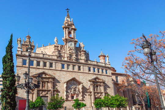 Baroque facade of historic Iglesia de los Santos Juanes in Valencia, Spain. Ornate sculptures and a clock tower rise above the bustling Plaza del Mercado near the central market