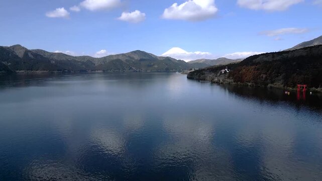 Wide panoramic view of Lake Ashi in Hakone surrounded by mountains under blue sky. Calm water with red torii gate visible on shore. Beautiful natural landscape in Kanagawa Prefecture Japan.
