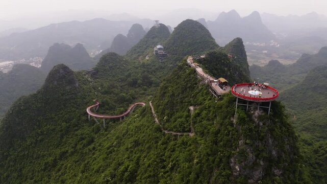 Gentle right-to-left drone orbit reveals the red circular observation deck and winding Skybridge elevated on lush karst cliffs in misty Yangshuo, Gulin. Hazy peaks, green trails, and distant ridges fr