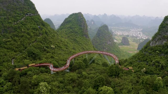Descending aerial view traces vibrant red Skybridge amid lush karst peaks in Yangshuo Gulin Guangxi. Hazy ridges and green valleys create serene scenic beauty for travel stock footage.