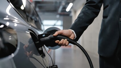 Obraz premium Captured in a dark, high-contrast close-up, a professional man's hand plugs a black charging cable into a sleek electric vehicle within an indoor parking garage.