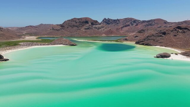 Aerial view of the turquoise waters meeting the white sands of Balandra Beach, contrasted against the rugged, dark mountains, La Paz, Baja California Sur, Mexico.