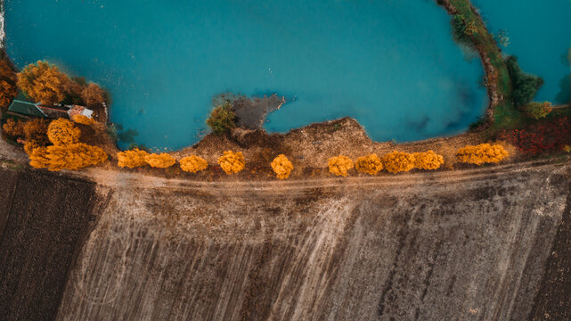 Aerial view of a tranquil, turquoise lake bordered by a vibrant strip of golden trees contrasting with the dark, earthy fields below, Novi Sad, Vojvodina, Serbia.