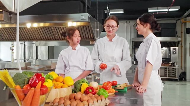 Three chefs in a commercial kitchen learn food prep as an instructor demonstrates tomatoes, surrounded by colorful vegetables and eggs on a stainless counter
