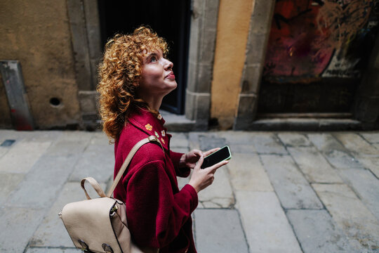 Curly haired woman holding smart phone and exploring alley