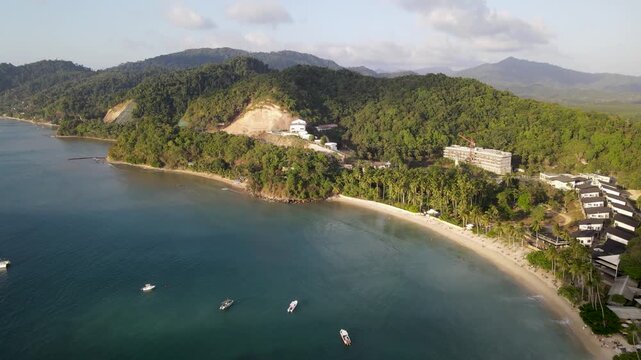 Aerial view of the coast, with turquoise waters meeting golden sands, lush green mountains, and buildings, El Nido, Mimaropa, Philippines.