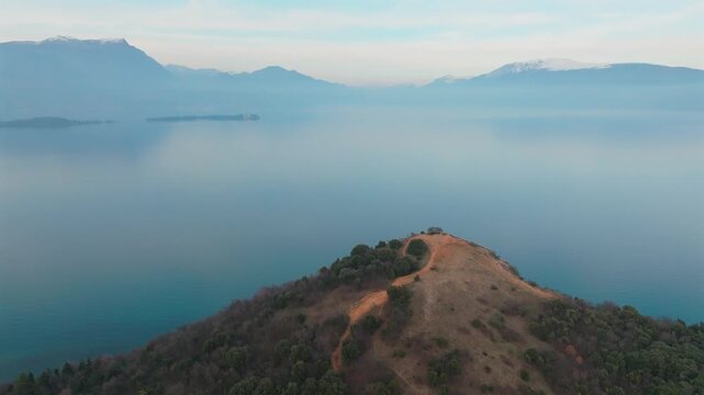 Aerial view of a hill covered with trees and grass, contrasts with the blue water of Lake Garda and snow-capped mountains, Manerba del Garda, Lombardia, Italy.
