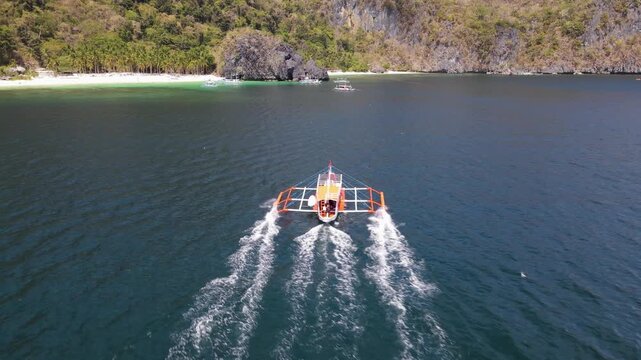 Aerial view of a boat sailing on the dark blue ocean near the shore with white sand and palm trees, El Nido, Mimaropa, Philippines.