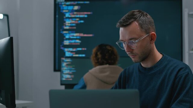 Focused diverse software engineers and AI developers working on programming code on computer screens in a dark office. Concept of machine learning, digital transformation, and future technology.