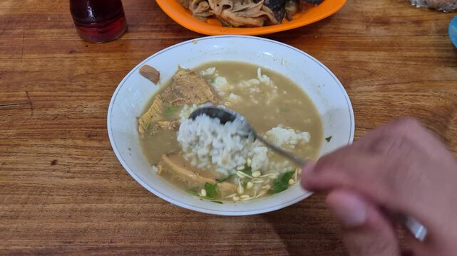 a bowl of traditional indonesian soup called soto daging containing sliced block of meat, rice, some spices, and broth  mixed with coconut milk while stirred using metal spoon.