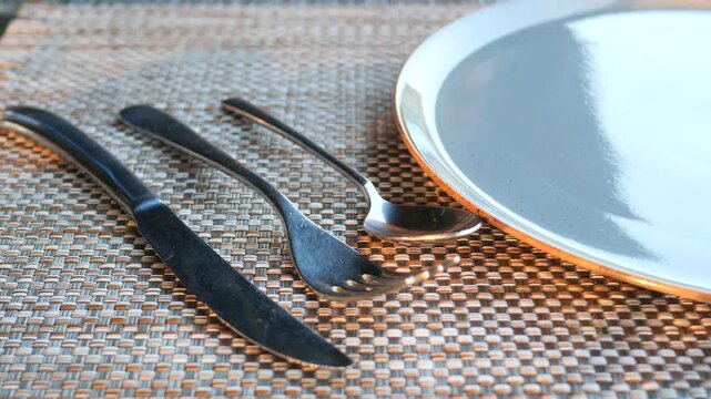 A close up view of an empty round ceramic dinner plate, spoon, fork, and knife, creating a neatly arranged table setting with a person picking up the cutlery in a dynamic, cinematic shot